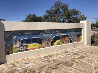 Tile mural measuring 18 ft wide and 3 ft 4 in tall inset into landscape wall. Mural depicts hand drawings of Mission Concepción, Mission San José, Mission San Juan, and Mission Espada. Watercolor brushstrokes of blues, orange and yellow are in the background and line drawings of plants and animals surround the missions. Four tile motifs from original San Jose Workshop tile patter run along the foreground.