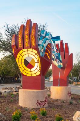 The sculpture, made of painted steel, is a pair of abstracted hands connected by a strand of rippling water. At the base of each hand, a mesquite leaf graphic is inscribed on the concrete base.